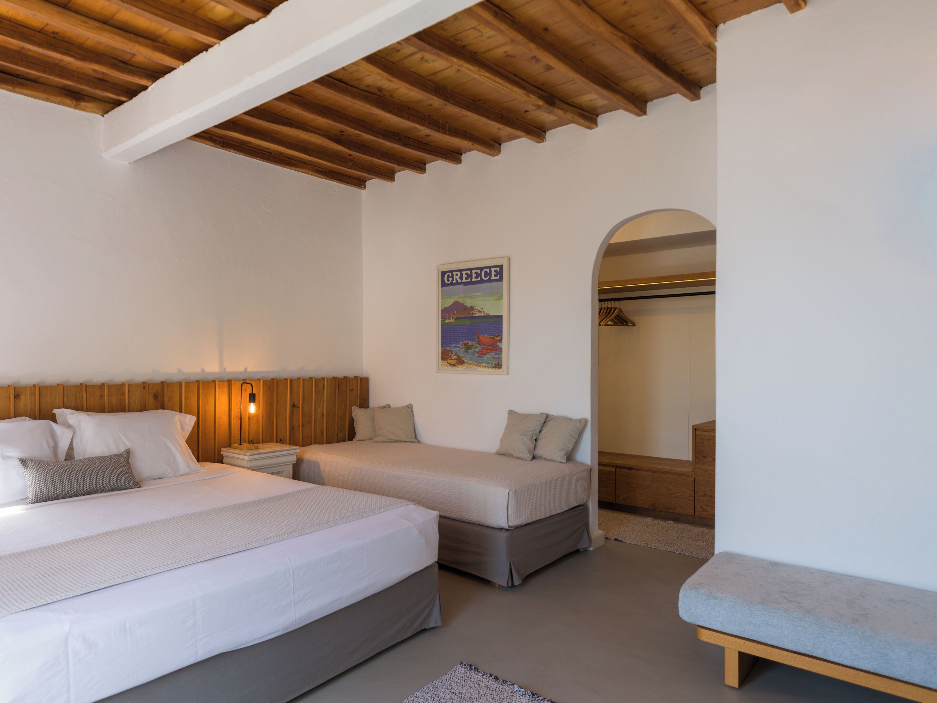A bedroom with two beds, a wooden ceiling, a poster of Greece, and a bench.
