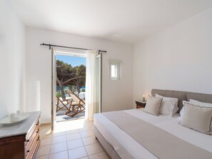 A bedroom with a bed, a nightstand, a dresser, a window with curtains, and a view of the outdoors. -  Rhenia Mykonos Hotel & Bungalows (Mykonos)