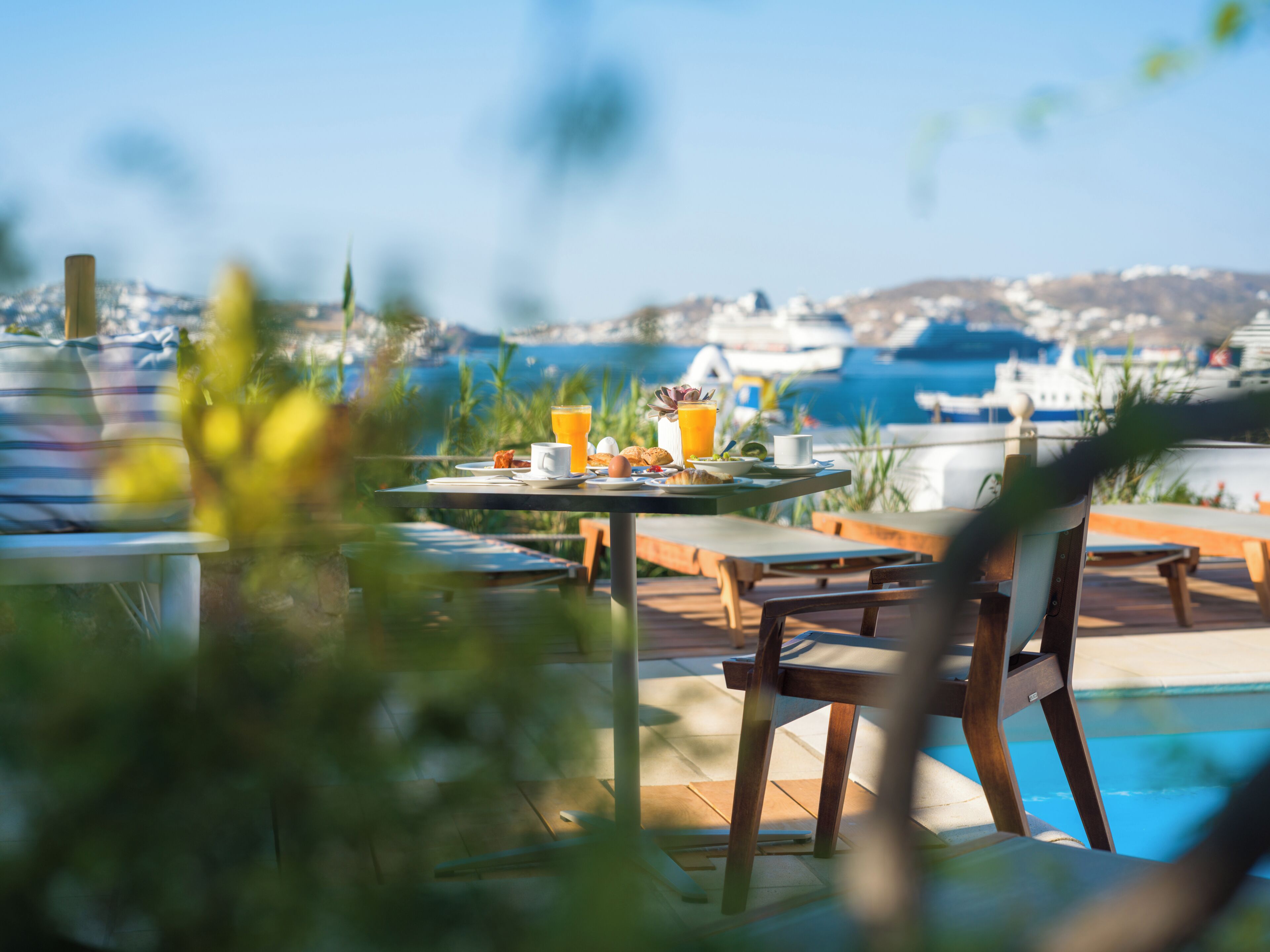 Un coin repas avec vue sur la mer et les bateaux, comprenant une table où sont disposés des aliments pour le petit-déjeuner et une piscine en arrière-plan.