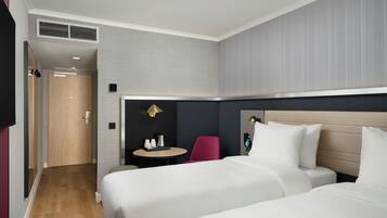 A hotel room with two beds, a small table with a lamp, a chair, and a door leading to another room.