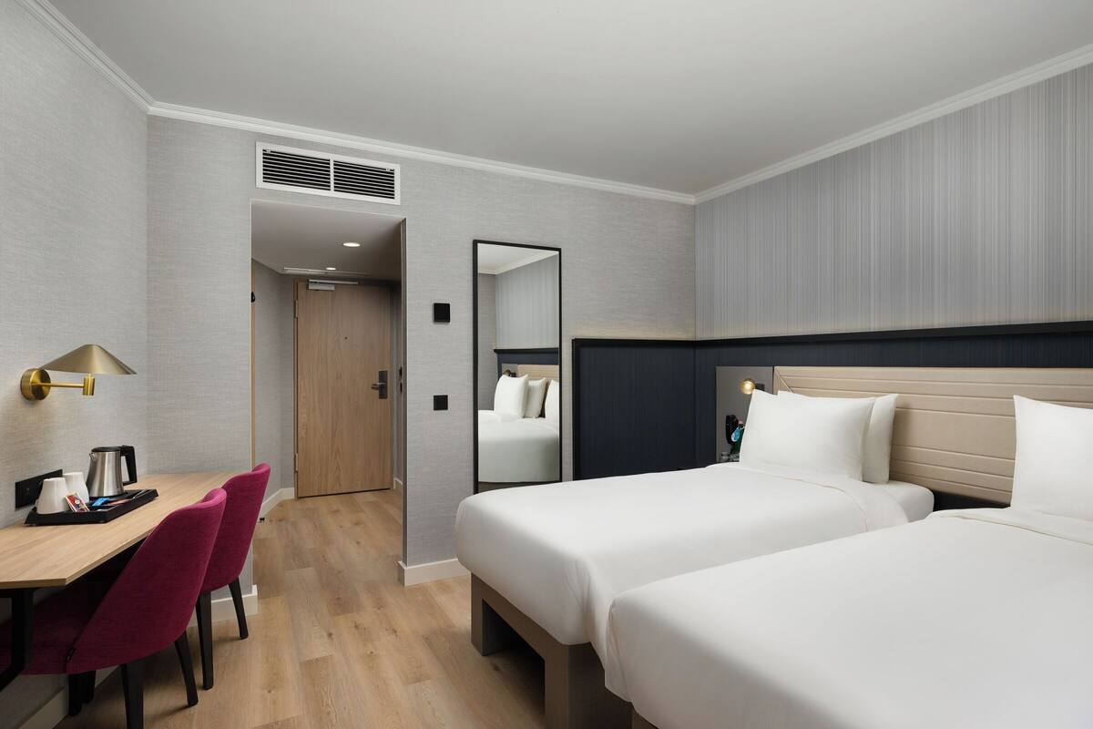 Habitación de hotel con dos camas, un escritorio con una silla, un espejo y una puerta que da a otra habitación.