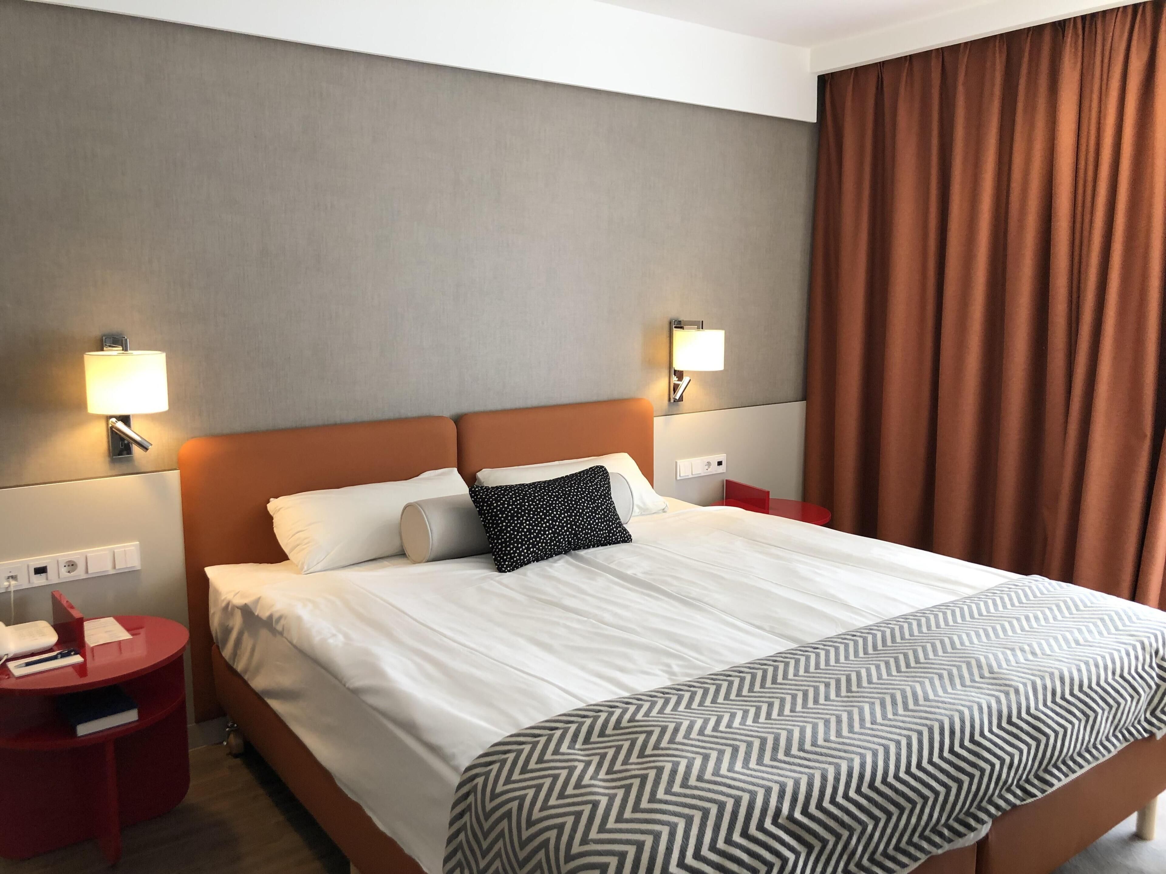 A hotel room with a large bed, two bedside tables, a red side table, and a wall-mounted lamp.