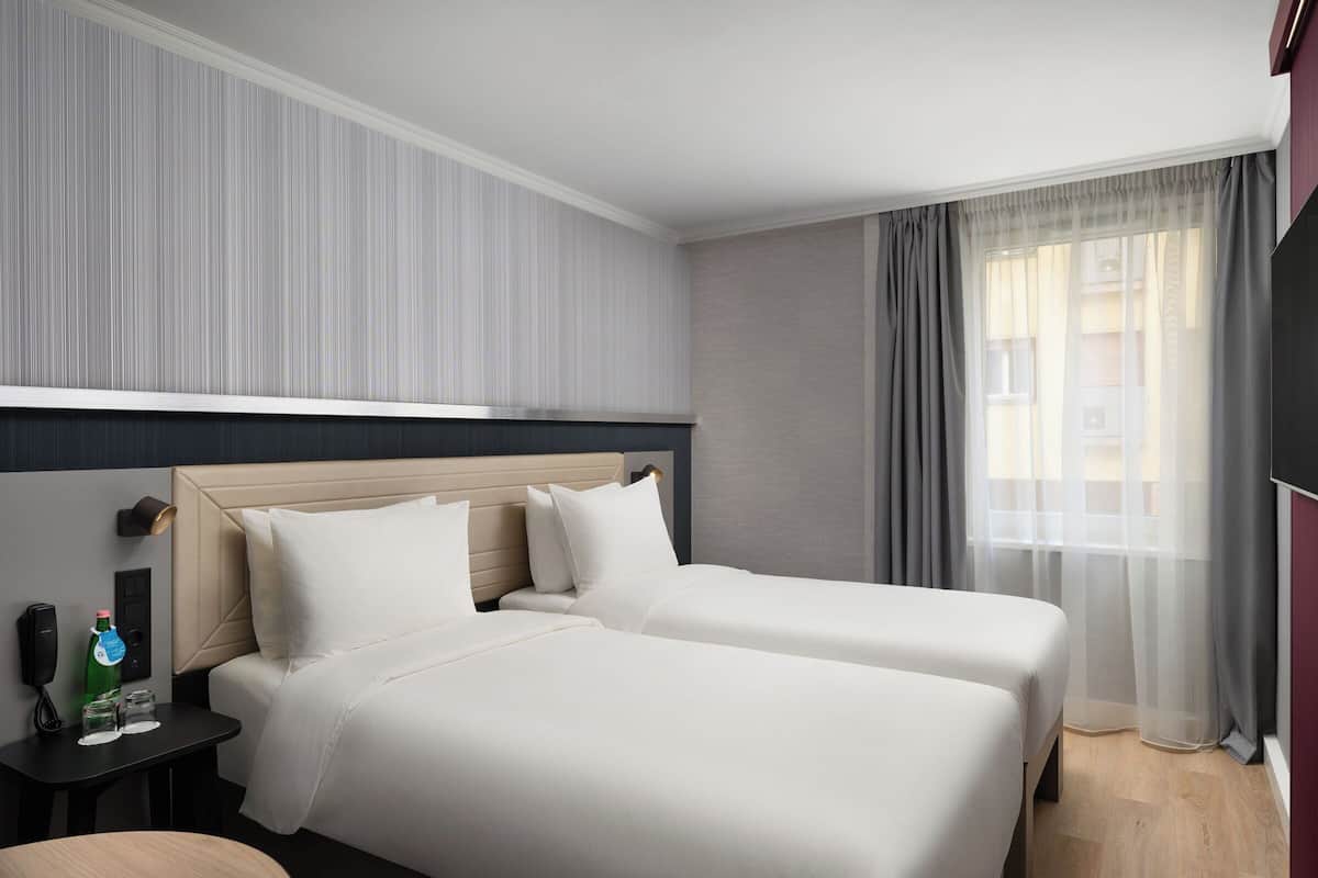 Habitación de hotel con dos camas, una mesita de noche con un teléfono y una botella, y una ventana con cortinas.