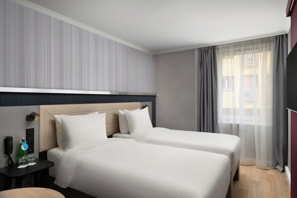 Habitación de hotel con dos camas, una mesita de noche con un teléfono y una botella, y una ventana con cortinas.