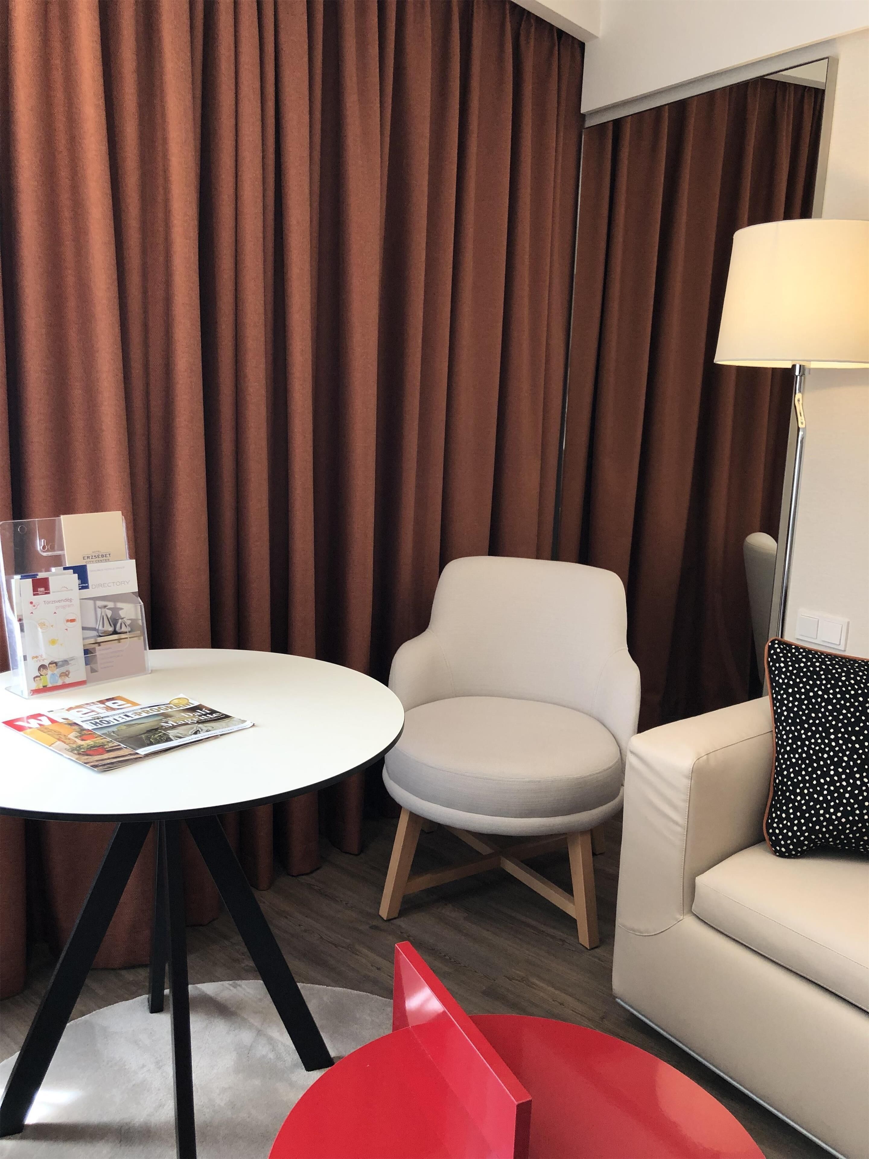 A room with a white round table, a beige armchair, a red accent chair, a white sofa, and a floor lamp.