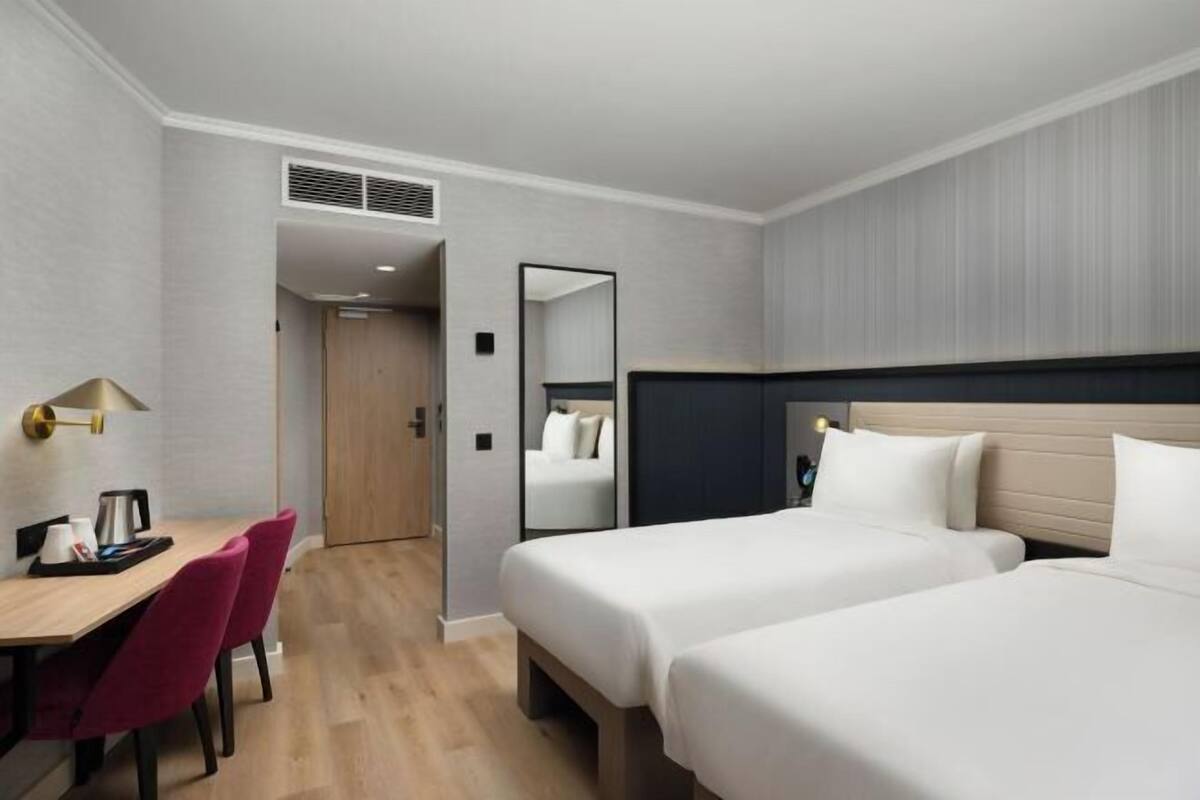 A hotel room with two beds, a desk with a chair, a mirror, and a door leading to another room.