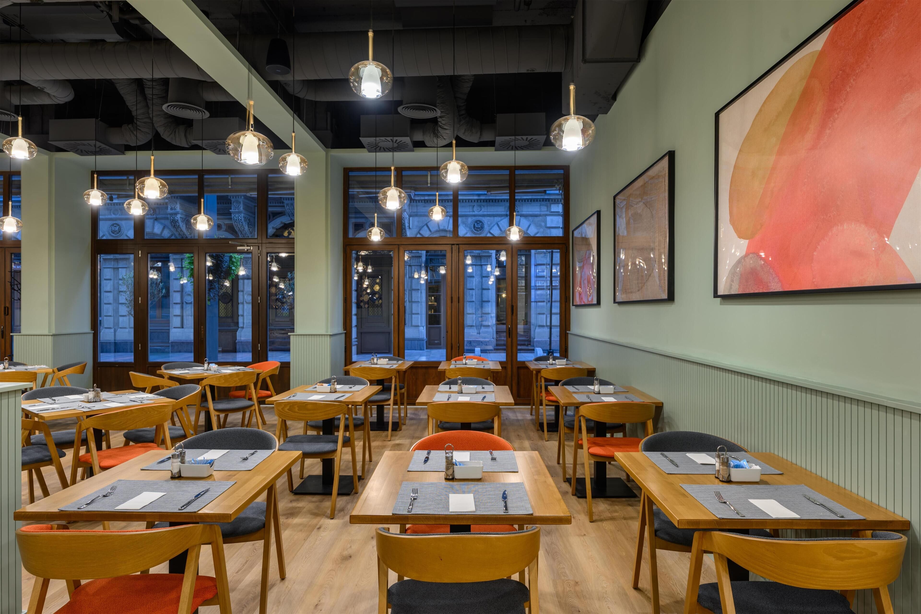 A restaurant interior with wooden tables and chairs, pendant lighting, large windows, and artwork on the walls.
