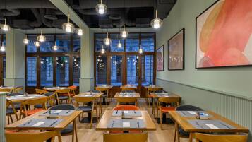 A restaurant interior with wooden tables and chairs, pendant lighting, large windows, and artwork on the walls.