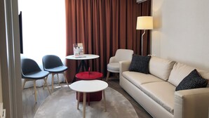A modern hotel room with a beige sofa, two armchairs, a small round table, and a red ottoman. A large window with brown curtains is visible, and a floor lamp is placed against the wall. - Hotel Erzsébet City Center Budapest, a member of Radisson Individuals (Budapest)