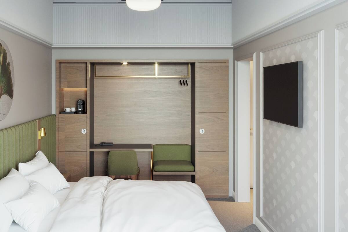 A hotel room with a bed, a desk with a chair, a TV, and a wall-mounted shelf.
