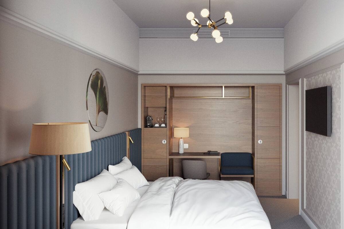 A modern hotel room with a large bed, a desk, and a chair. There is a wardrobe, a TV, and a lamp.