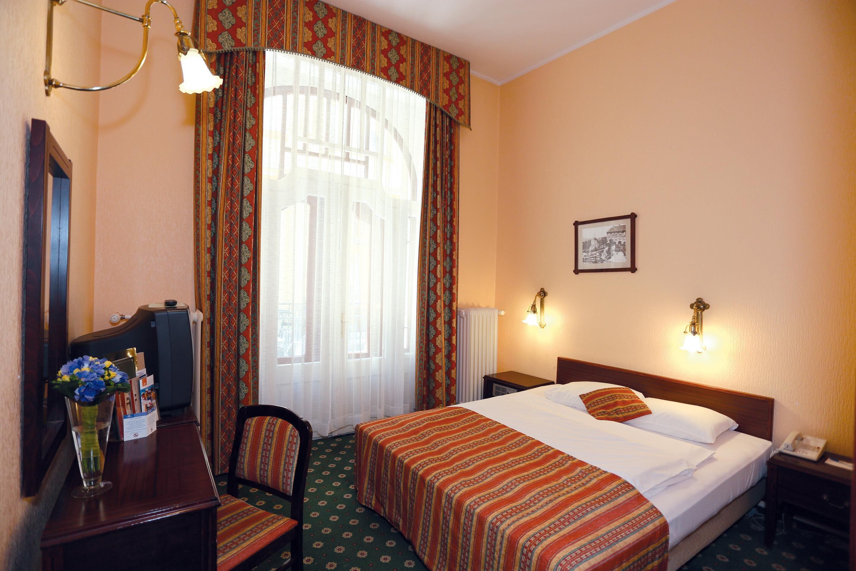 Une chambre d’hôtel avec un lit, un bureau avec une lampe, une chaise et une fenêtre avec des rideaux.