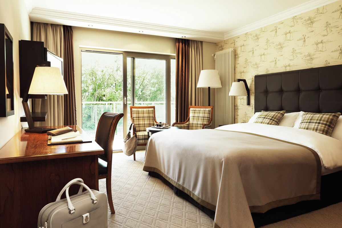 A hotel room with a large bed, a desk with a lamp, a chair, and a suitcase.