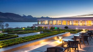 Een modern hotel met een terras dat uitkijkt over een waterpartij en bergen.