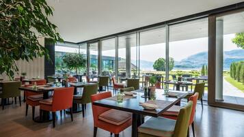 A modern restaurant with large windows offering a view of a lake and mountains. Tables are arranged with chairs, and there are potted plants and glassware on the tables.