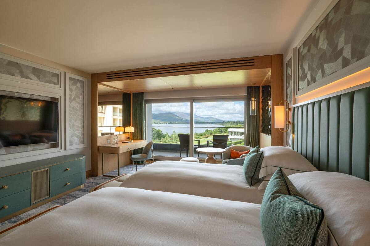 A hotel room with two large beds, a desk, and a chair. There is a large window offering a view of the mountains and a lake.