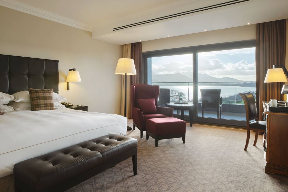 A hotel room with a large bed, a red armchair, a wooden desk, and a view of a body of water through large windows.