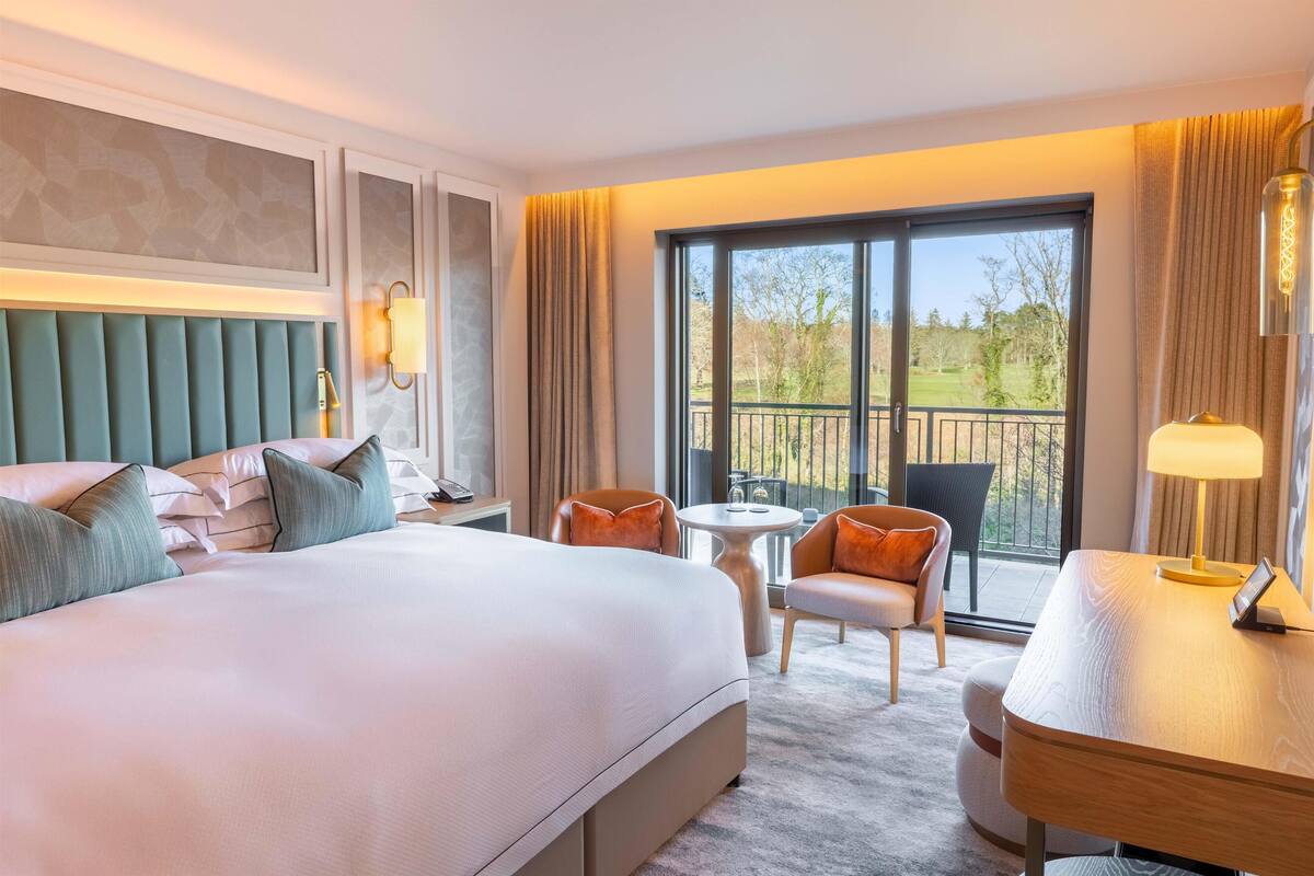 A hotel room with a large bed, a desk, two chairs, a small table, and a balcony with a view of trees and a building.