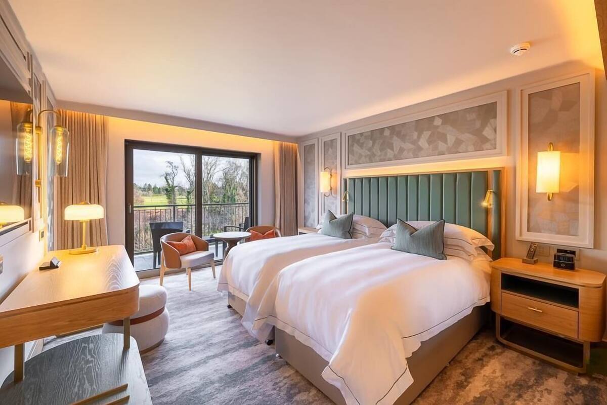 A hotel room with two beds, a desk, a chair, a small table, and a balcony with a view of the outdoors.