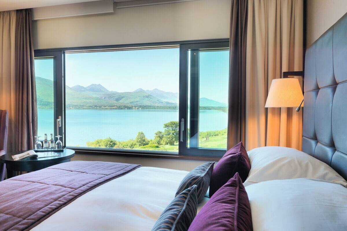 A hotel room with a large bed, a nightstand, a lamp, and a view of a lake and mountains.