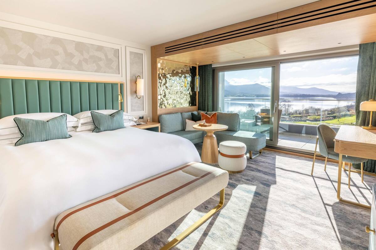 A modern hotel room with a large bed, a sofa, a dining area, and a view of a lake and mountains.
