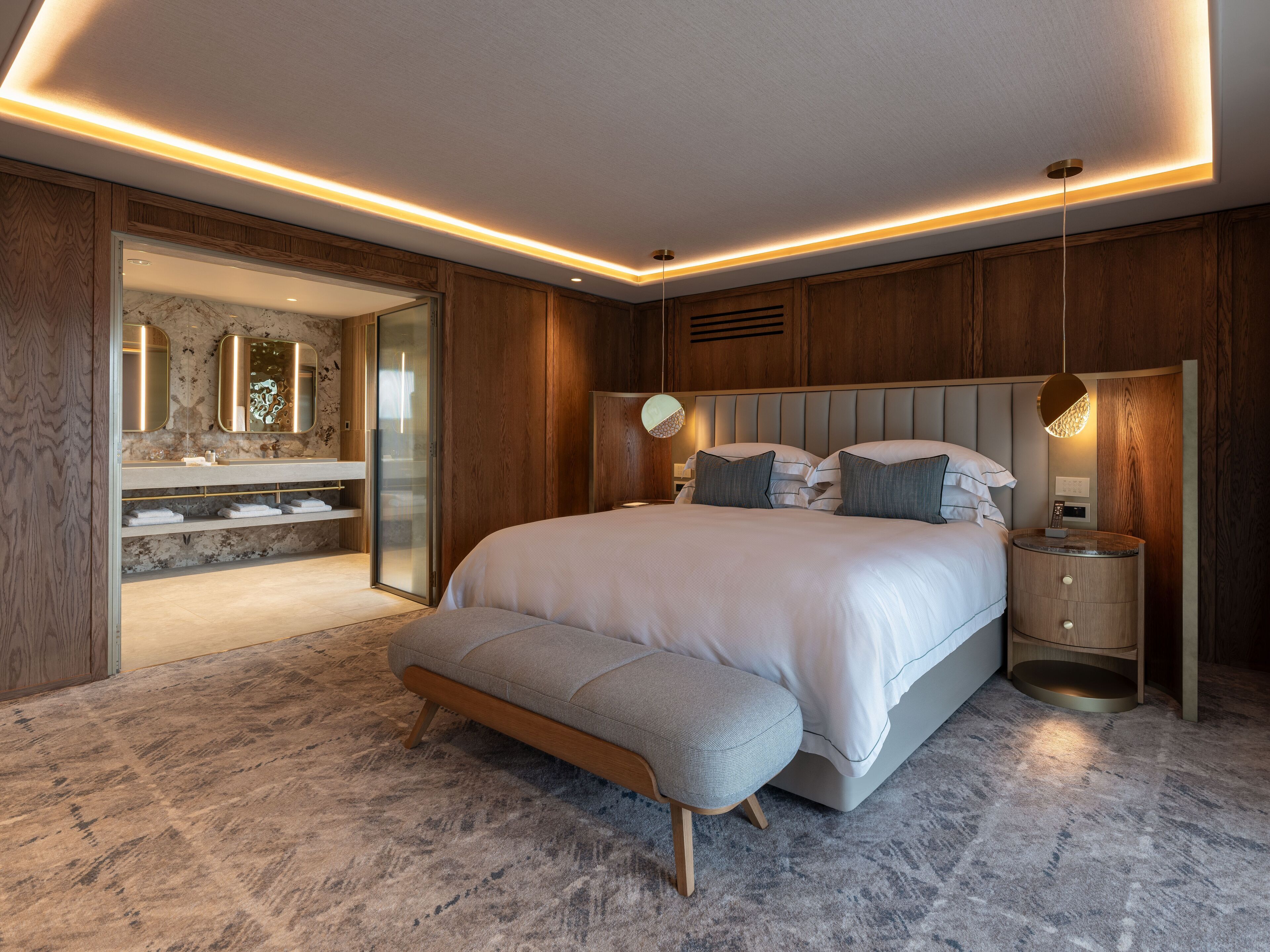 Un dormitorio moderno con una cama grande, mesitas de noche, un banco y un baño con tocador y espejo.