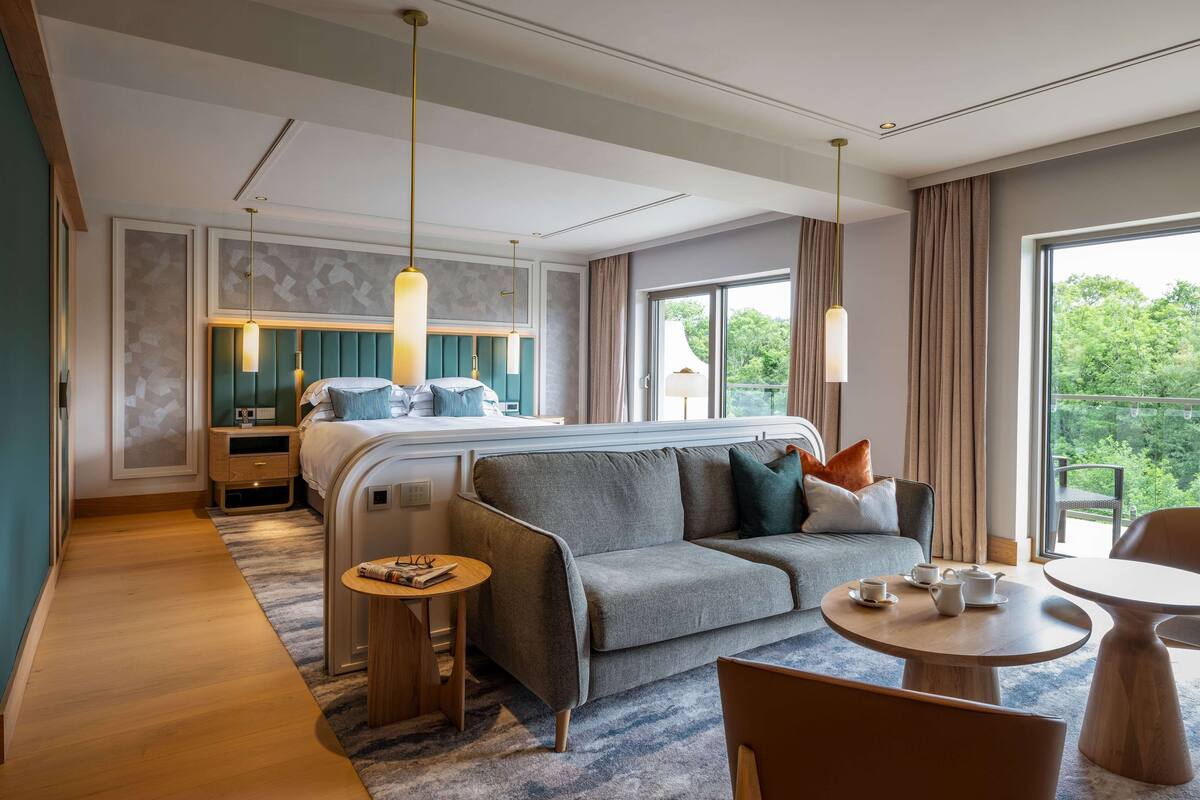 A modern hotel room with a large bed, a sofa, a small table, and a balcony with a view of the greenery.