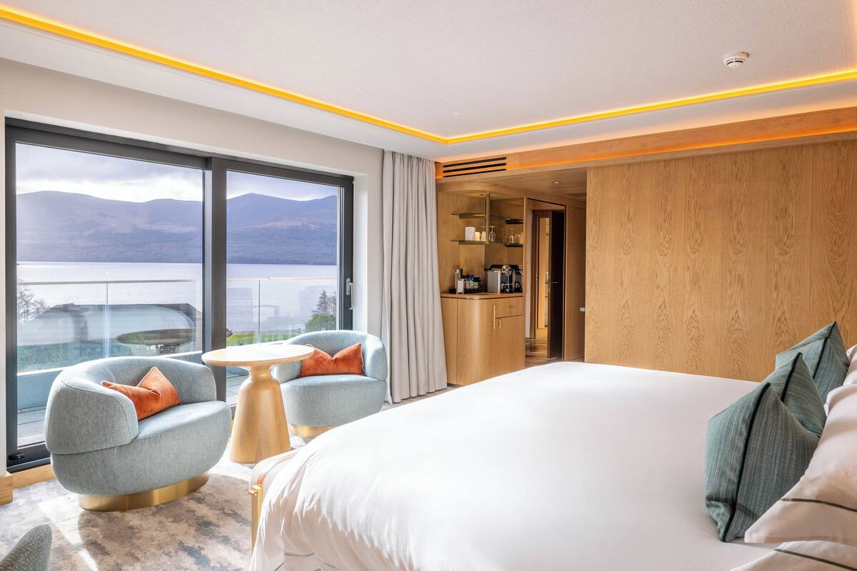A modern hotel room with a large bed, a seating area with a table, and a view of mountains through large windows.