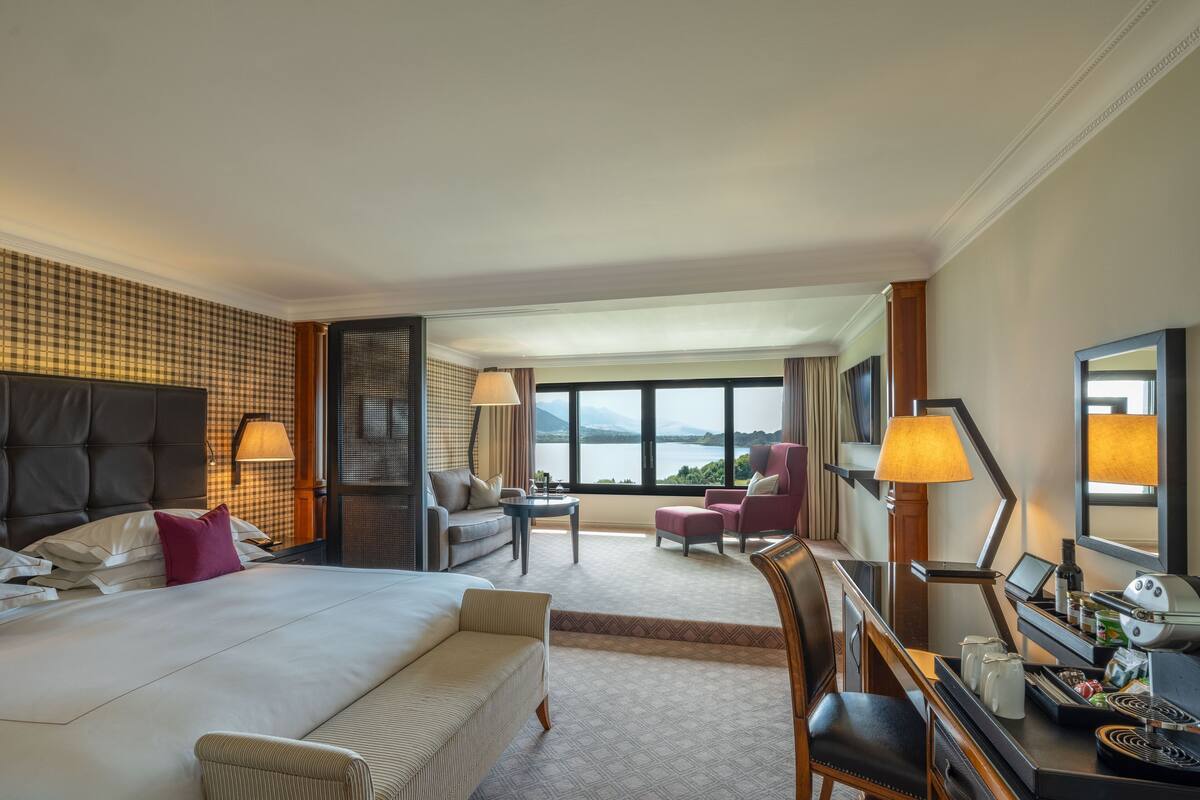 A hotel room with a large bed, a desk with a computer, a chair, a sofa, and a view of a lake.
