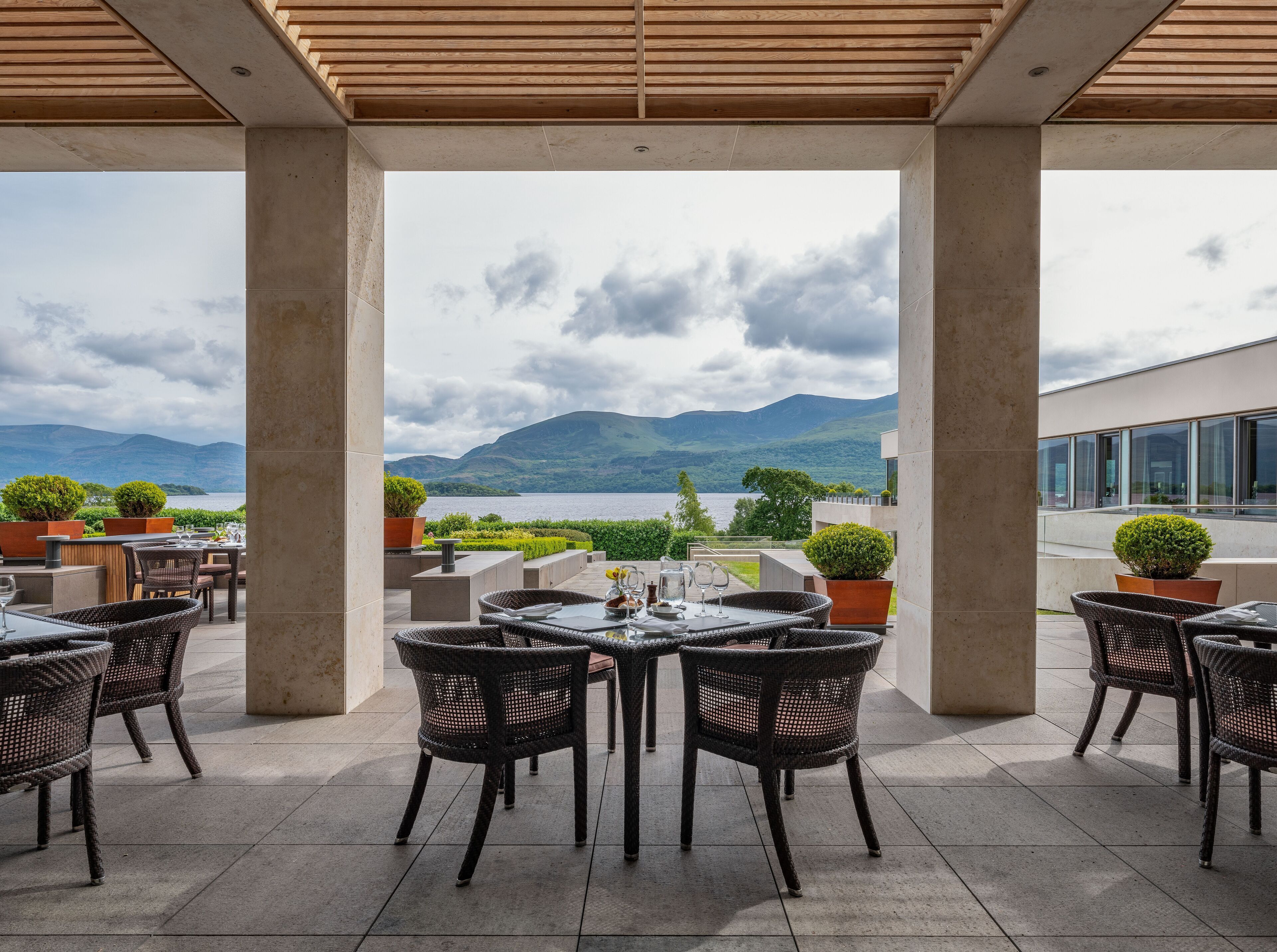 Un patio al aire libre con mobiliario de mimbre y una mesa lista para comer, con vistas a un lago y montañas.