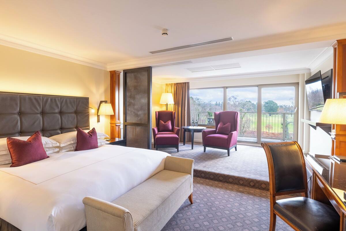 A hotel room with a large bed, two armchairs, a small table, a TV, and a balcony with a view.