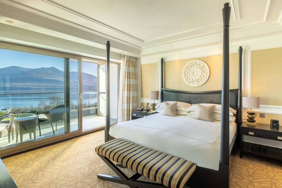 A hotel room with a large bed, a bench, a nightstand, a lamp, a decorative wall piece, and a view of mountains and water through large windows.
