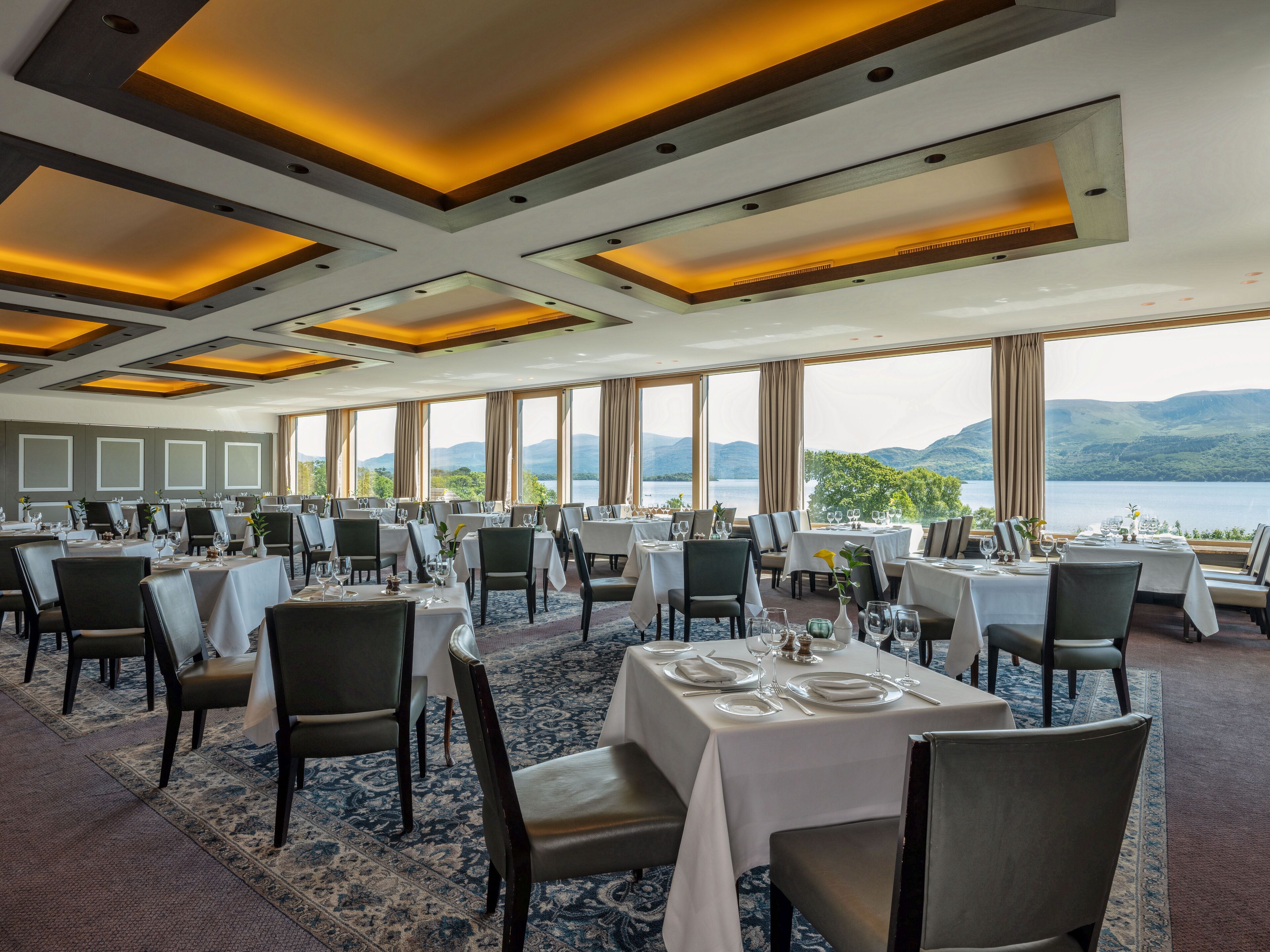 Ein Restaurant mit mehreren Esstischen, große Fenster mit Blick auf einen See und Berge sowie ein modernes Decken­design mit Einbaustrahlern.