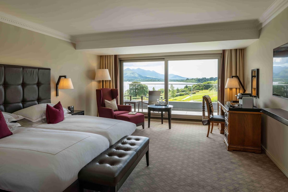 A hotel room with a large bed, a desk, a chair, and a view of a lake and mountains.