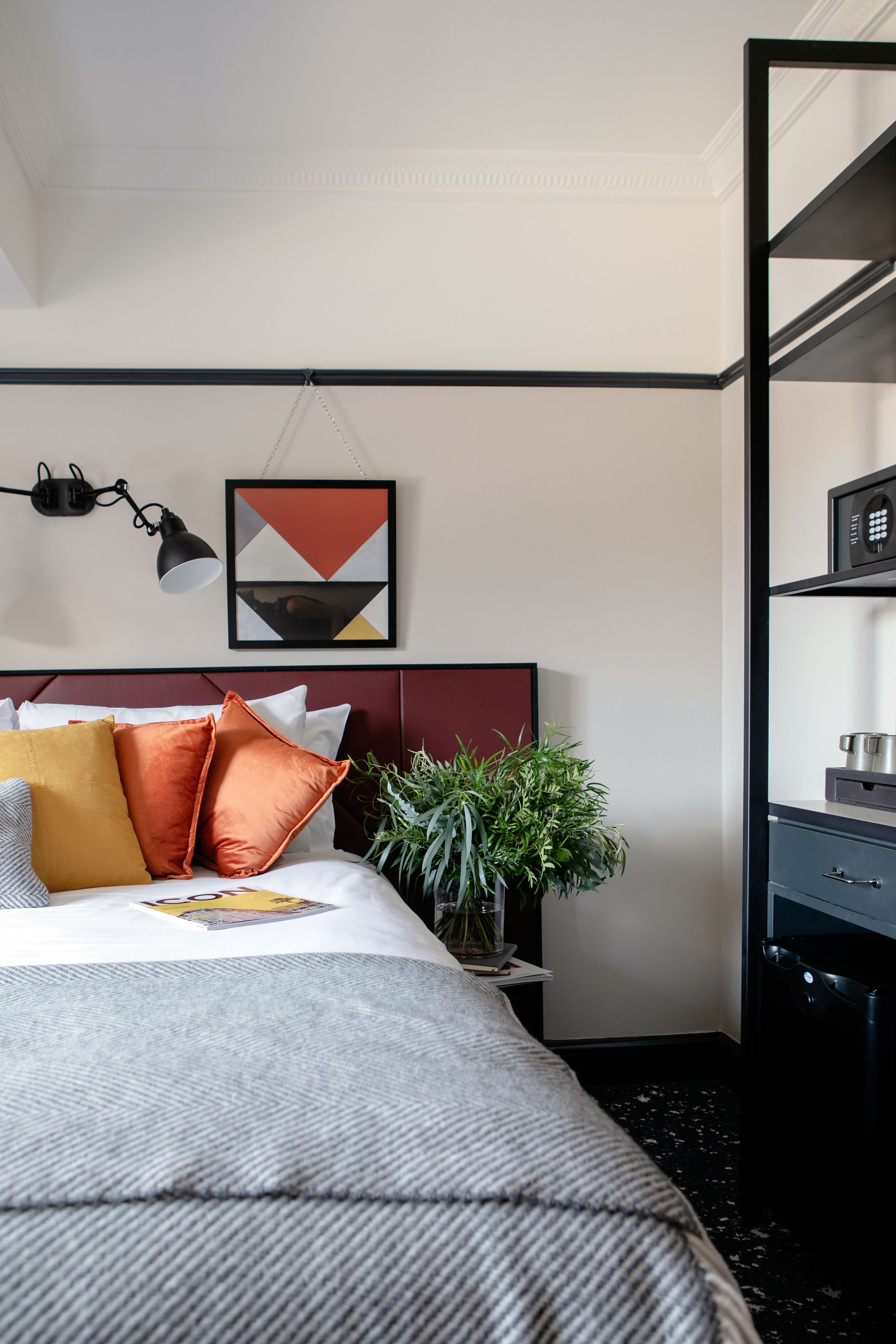 Un dormitorio ordenado con cama, almohadas, una planta en la mesita de noche y una repisa con un hervidor.