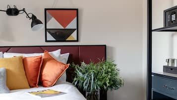 Un dormitorio ordenado con cama, almohadas, una planta en la mesita de noche y una repisa con un hervidor.