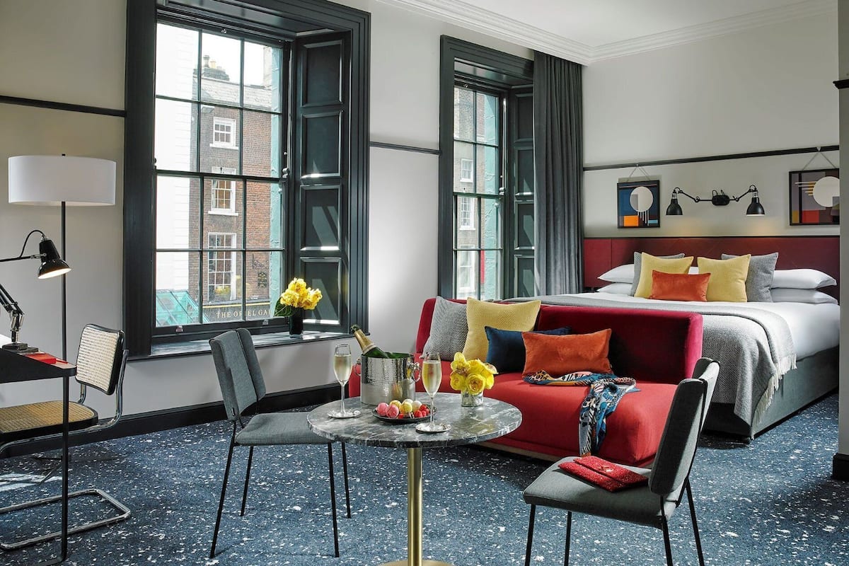 Quarto de hotel com cama, escrivaninha com cadeira, mesa redonda com um vaso de flores e janelas amplas.
