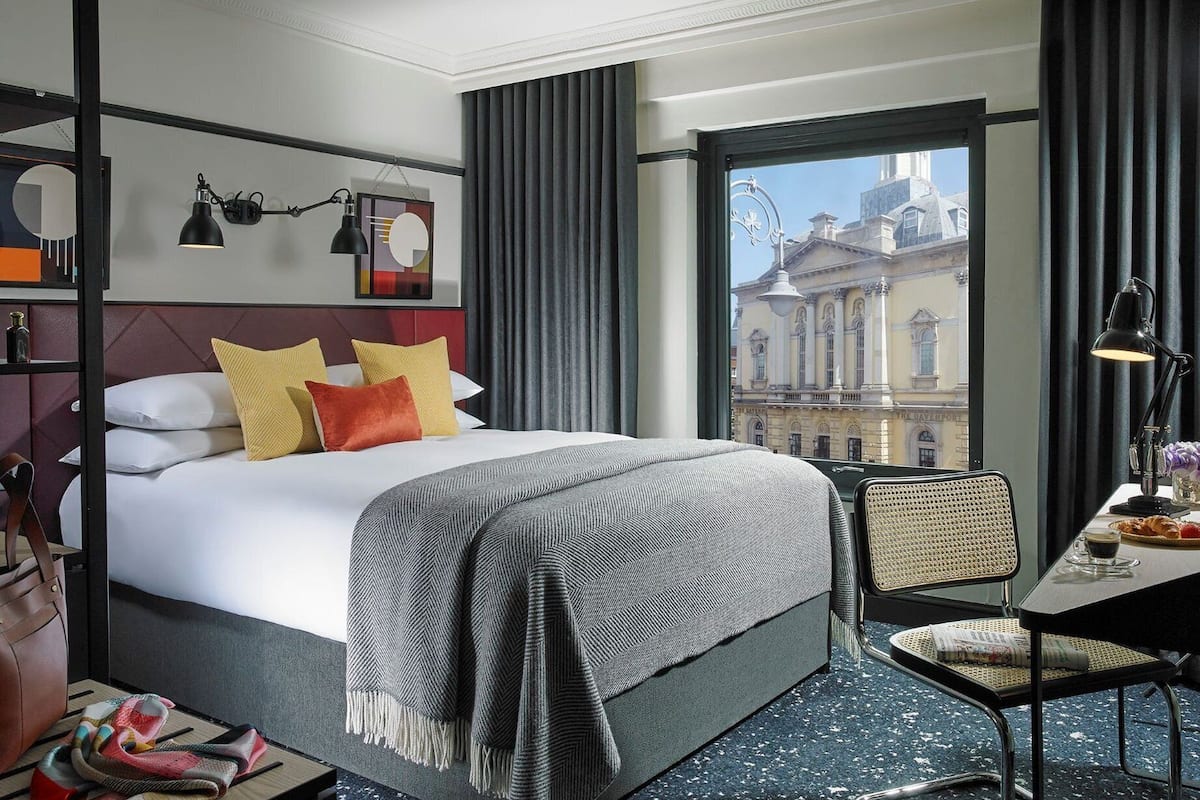 Quarto de hotel com uma cama grande, uma mesa com cadeira, vista para um edifício histórico e uma janela com cortinas.