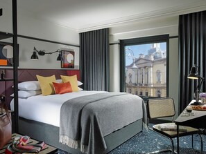 Habitación de hotel con una cama grande, un escritorio con silla, vista a un edificio histórico y una ventana con cortinas.