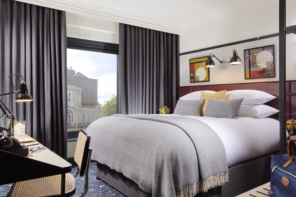 Quarto de hotel com uma cama grande, uma escrivaninha com luminária, uma cadeira e vista para a cidade pela janela.
