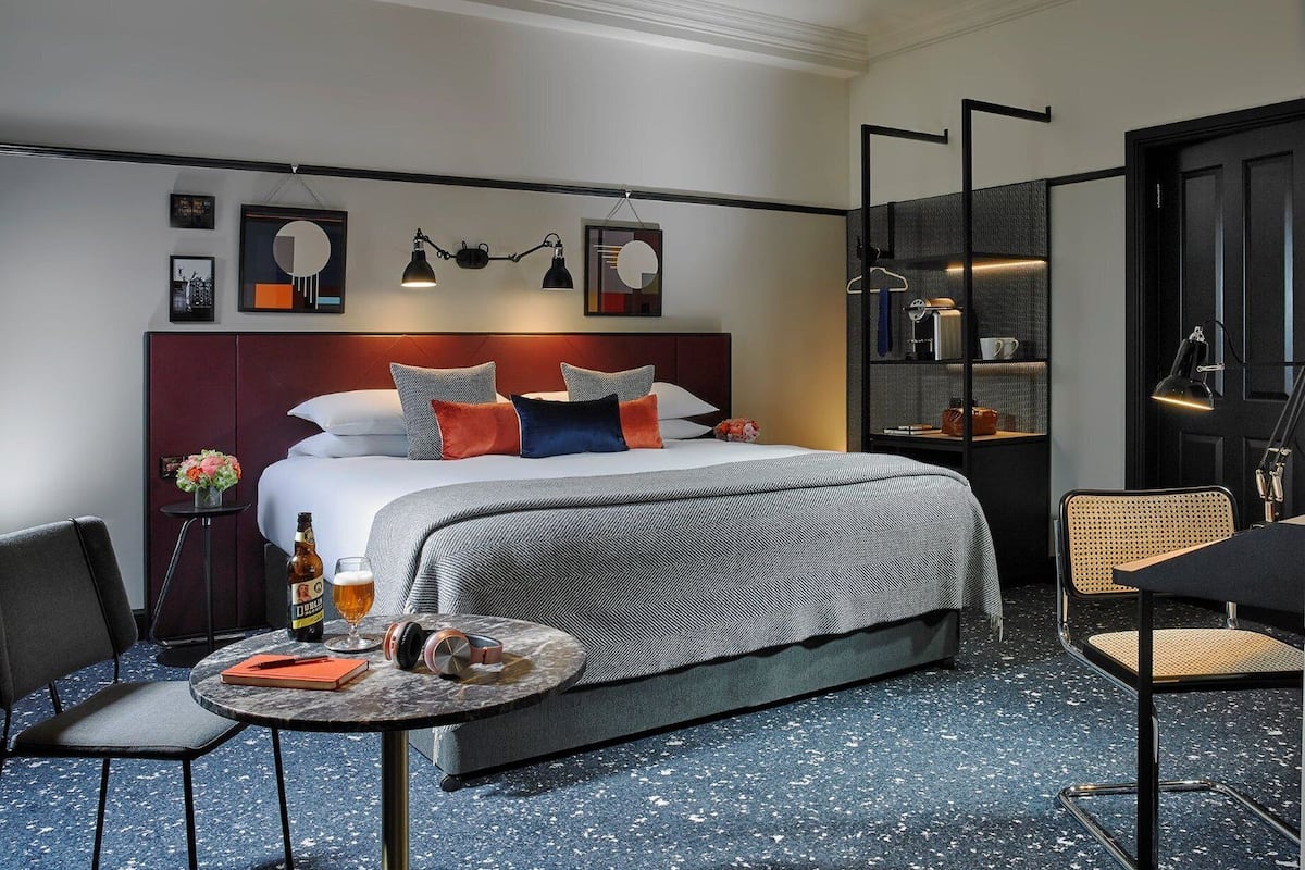 Quarto de hotel moderno com uma cama grande, uma mesa de centro redonda, uma cadeira e uma estante de livros.