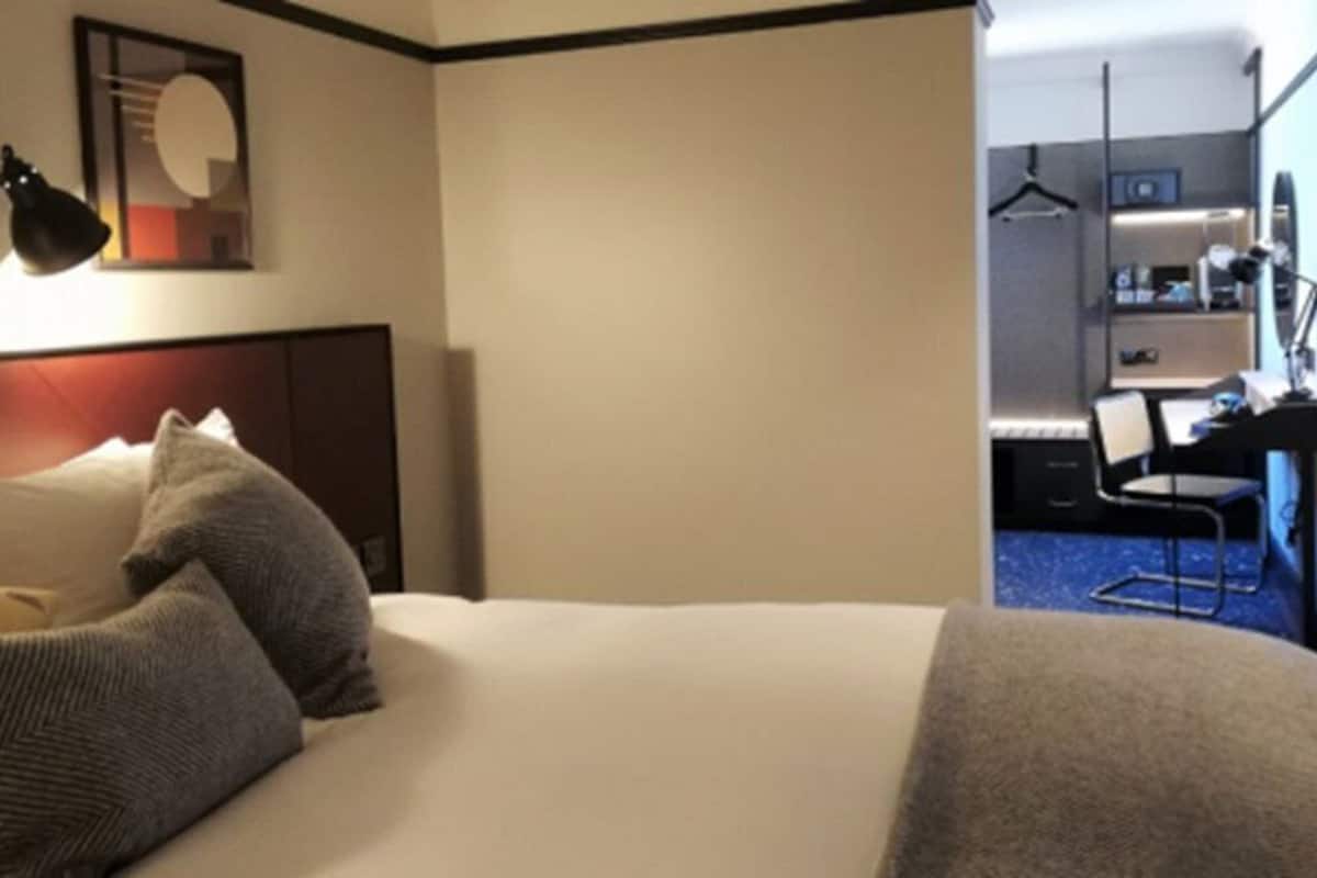 Quarto de hotel com cama, almofadas, escrivaninha com cadeira, luminária e espelho.