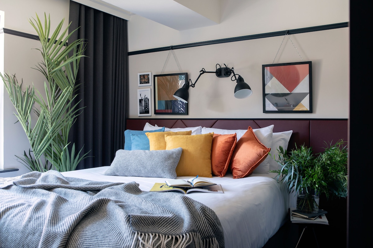A neatly made bed with a book, a potted plant, and a wall-mounted lamp.