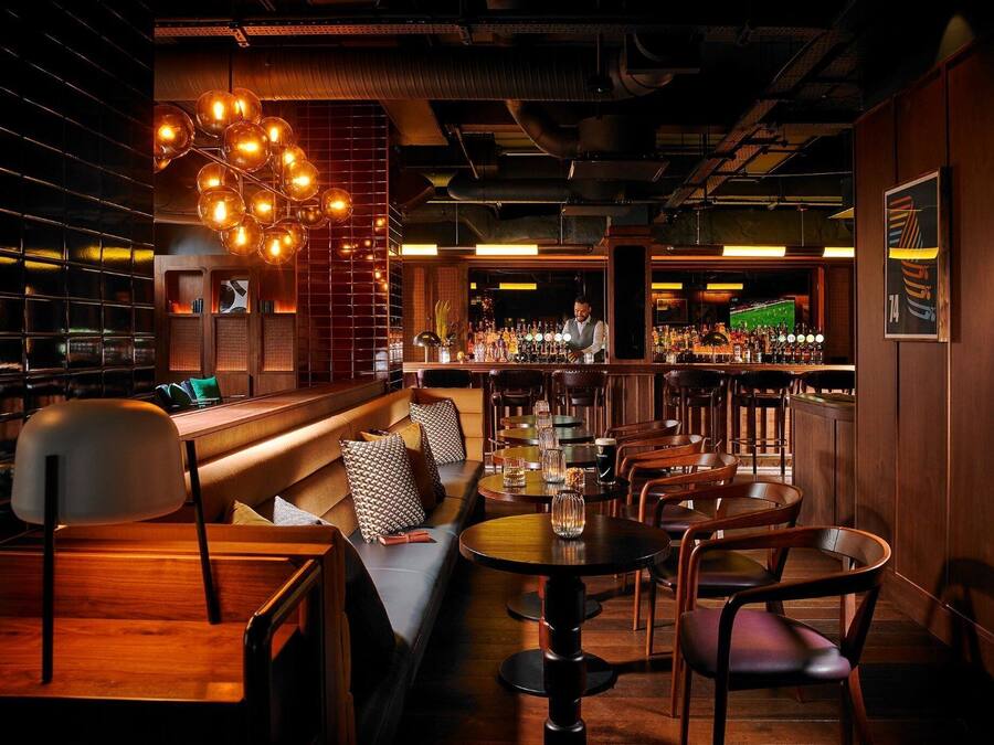 Una zona de bar con mobiliario de madera, un barman y una barra bien surtida.