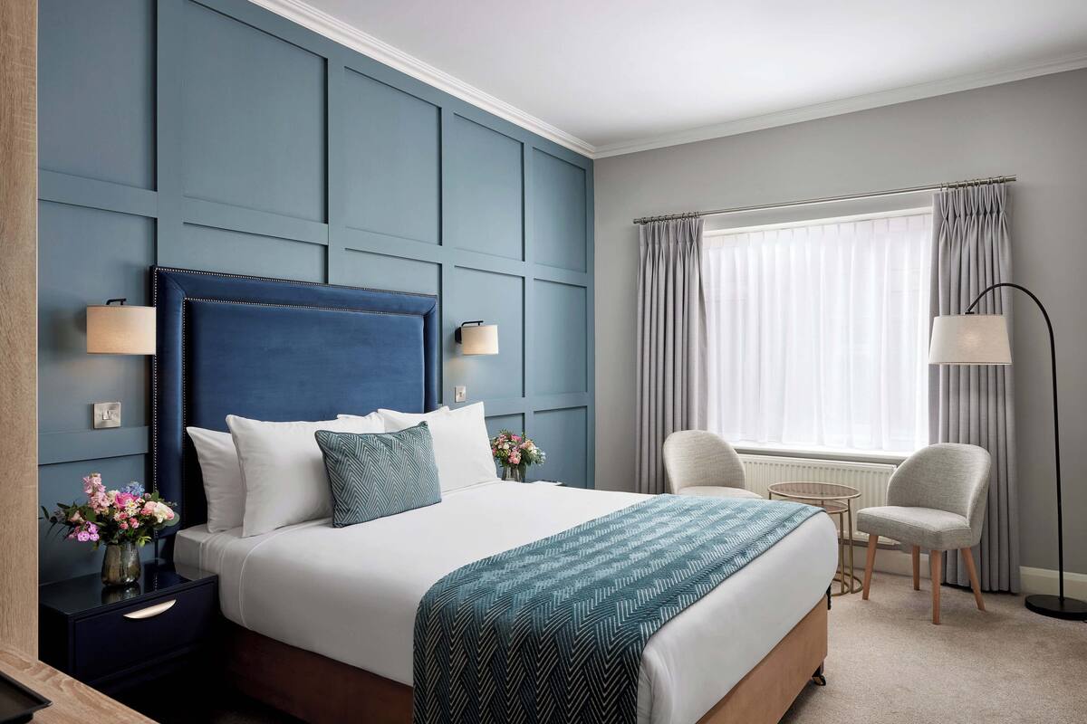 A hotel room with a large bed, a nightstand with a lamp, a chair, and a window with curtains.