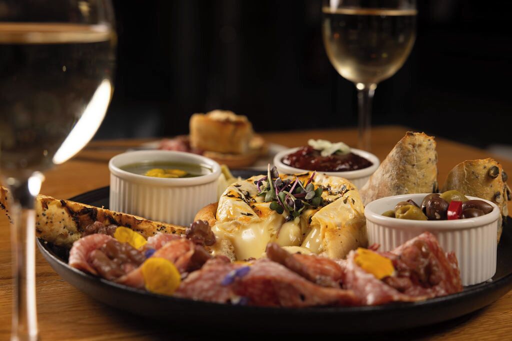 Una tabla de fiambres con diferentes embutidos, quesos y acompañamientos sobre un plato, junto a una copa de vino blanco.