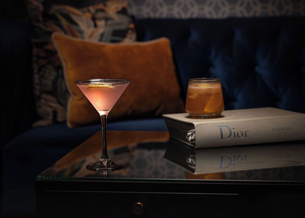 Una copa de martini sobre una superficie reflectante, con un libro titulado 'Dior' a su lado.