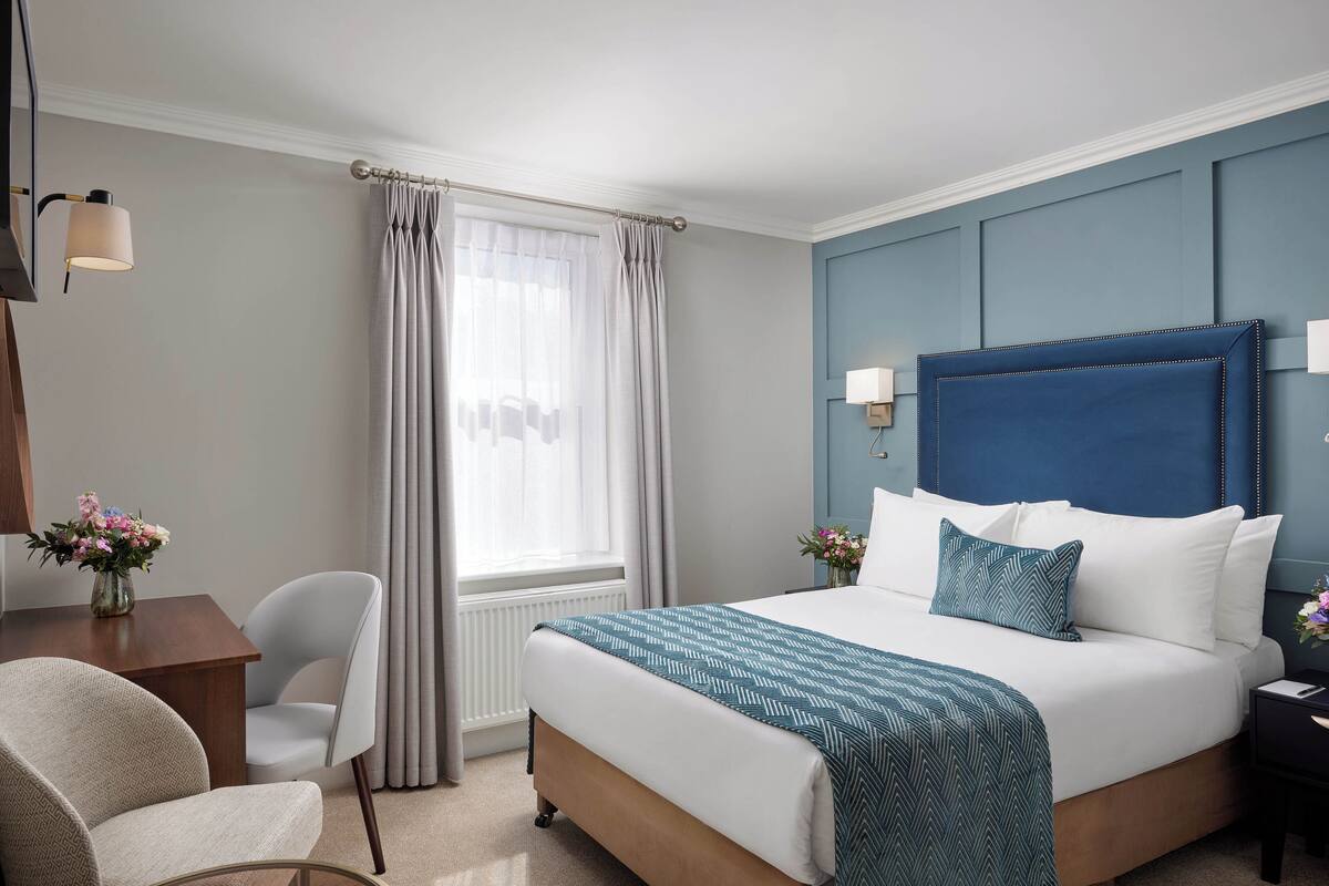 A hotel room with a large bed, a desk with a chair, a window with curtains, and a blue accent wall.