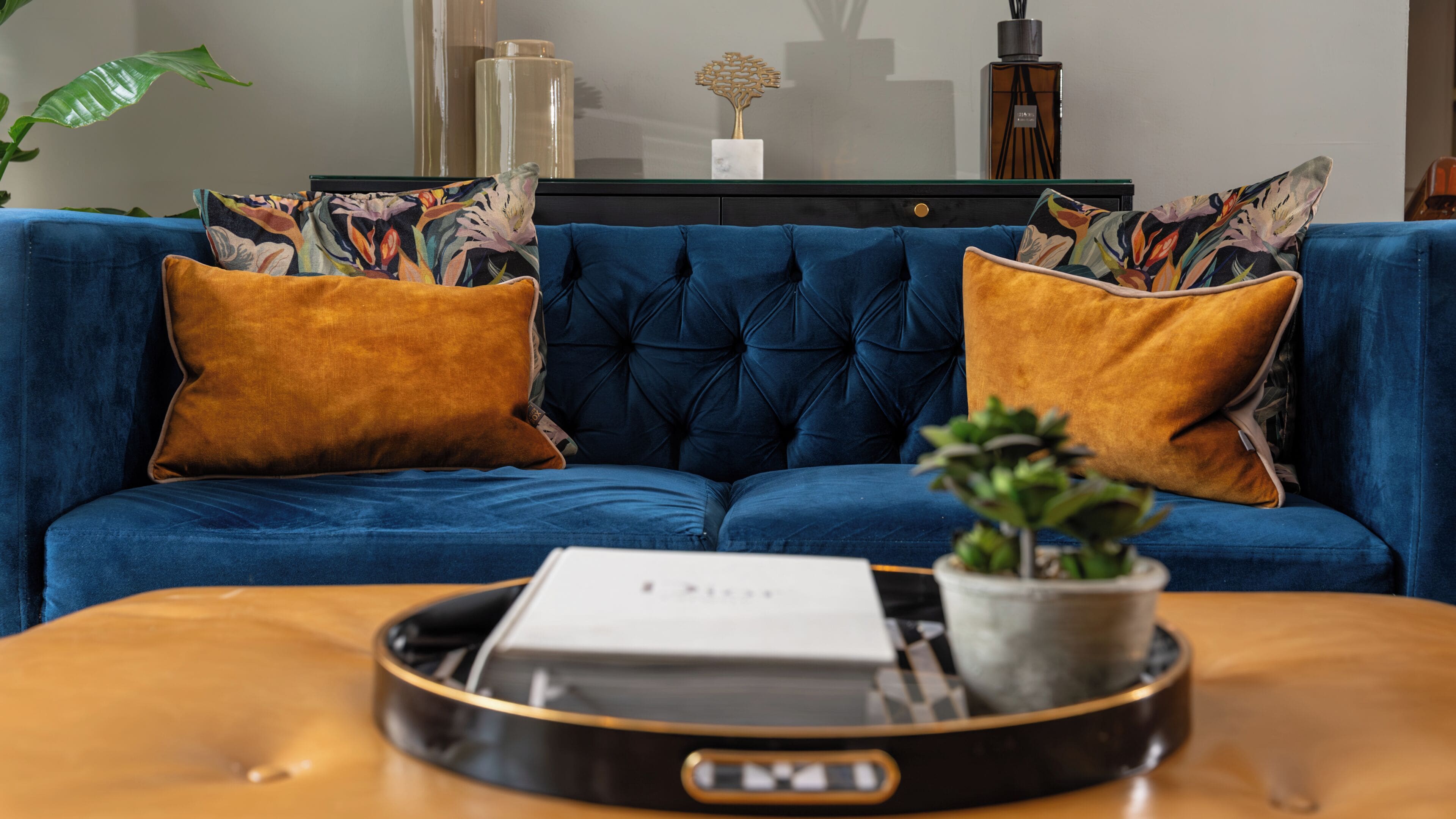 A blue sofa with two mustard-colored pillows, a small potted plant, and a tray with a white object on a wooden table.