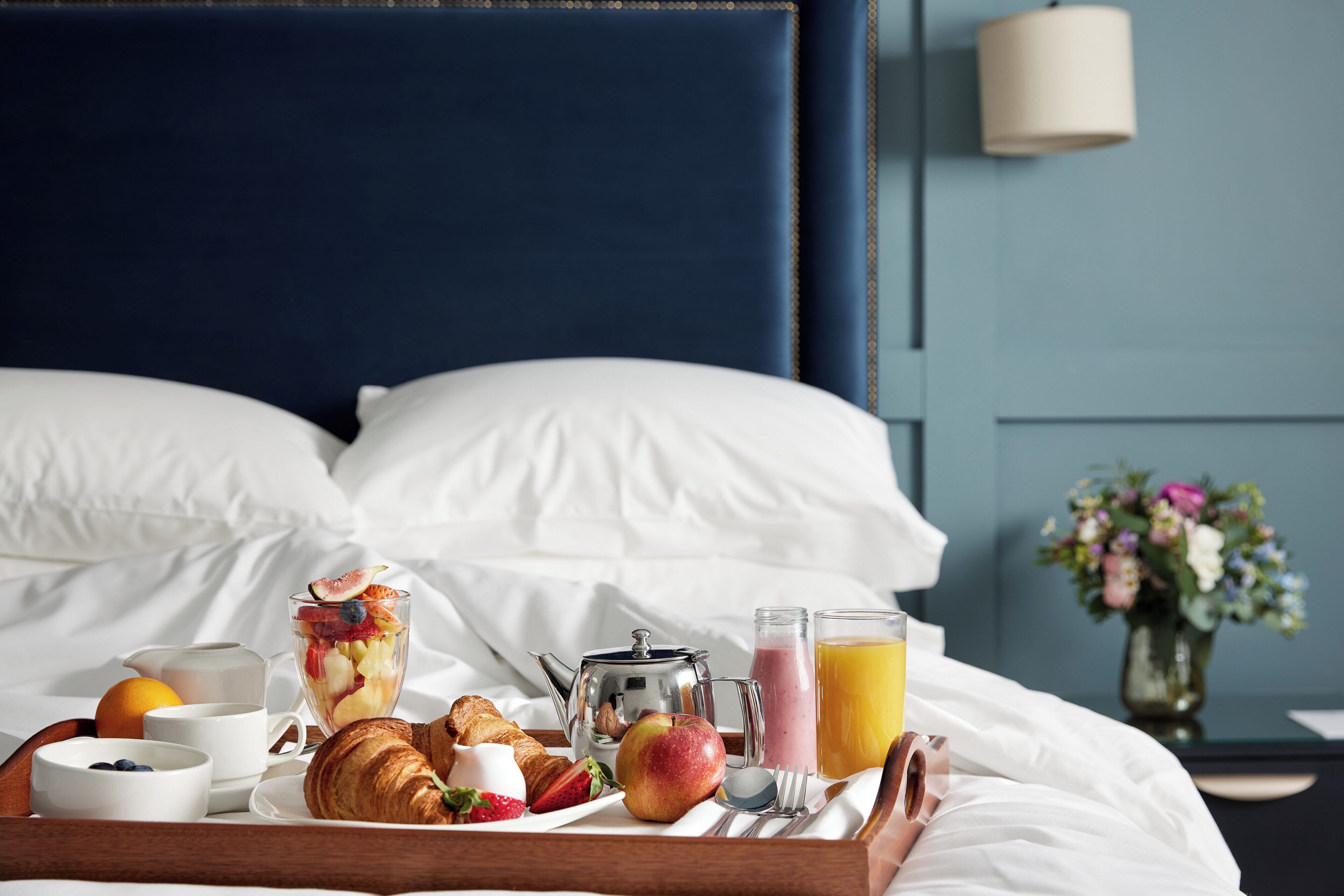 A neatly made breakfast tray on a bed with a variety of food items, a teapot, and a glass of orange juice.